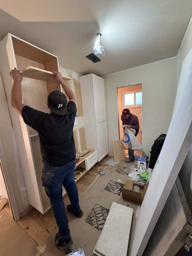 Two workers installing white cabinetry in a narrow room under renovation, one lifting an upper cabinet into place while the other handles materials near the doorway, with tools, boxes and protective floor covering around them.