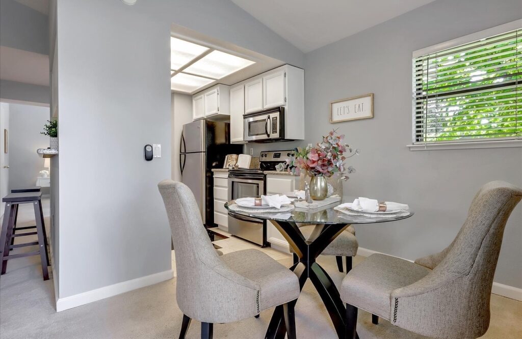 Dining area with a round glass table set for four and gray upholstered chairs, next to a window, opening into a kitchen with white cabinets and stainless steel appliances.