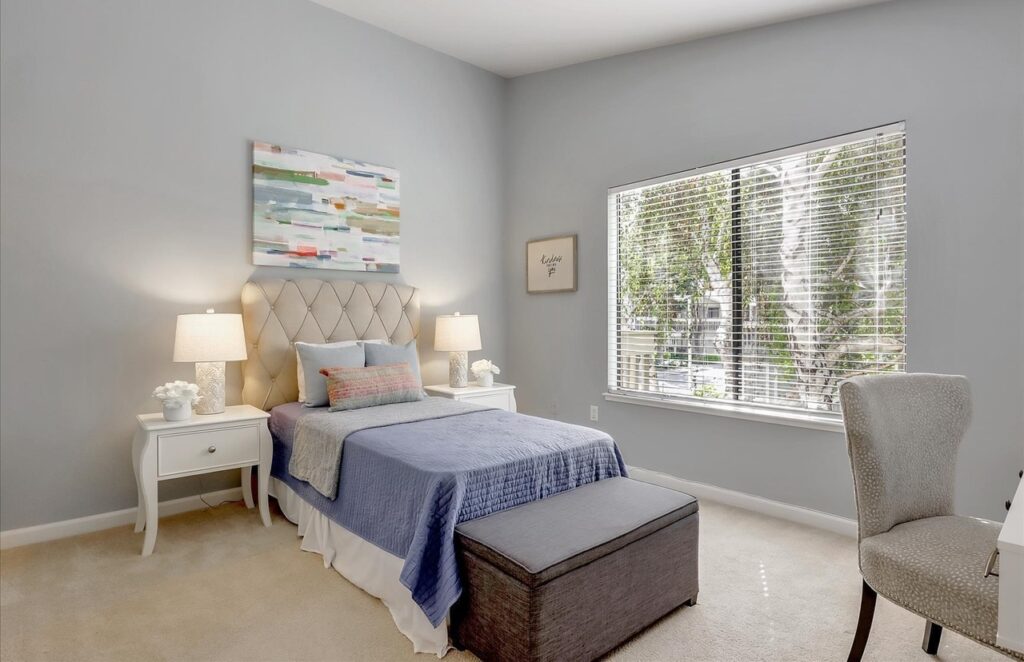 Carpeted bedroom with a bed with a tufted headboard and blue bedding, white nightstands with lamps, a storage bench, an upholstered chair and a large window with blinds overlooking trees.