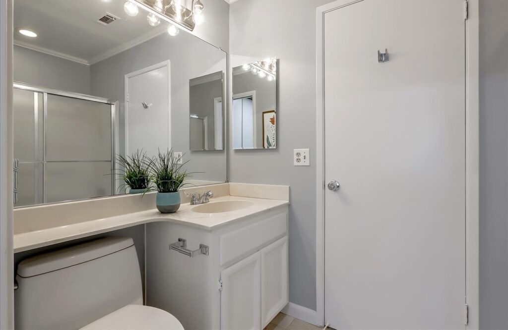 Bathroom with a white vanity and sink, a large wall mirror with a smaller mirrored cabinet, a toilet and a white door, with a glass-enclosed shower reflected in the mirror.