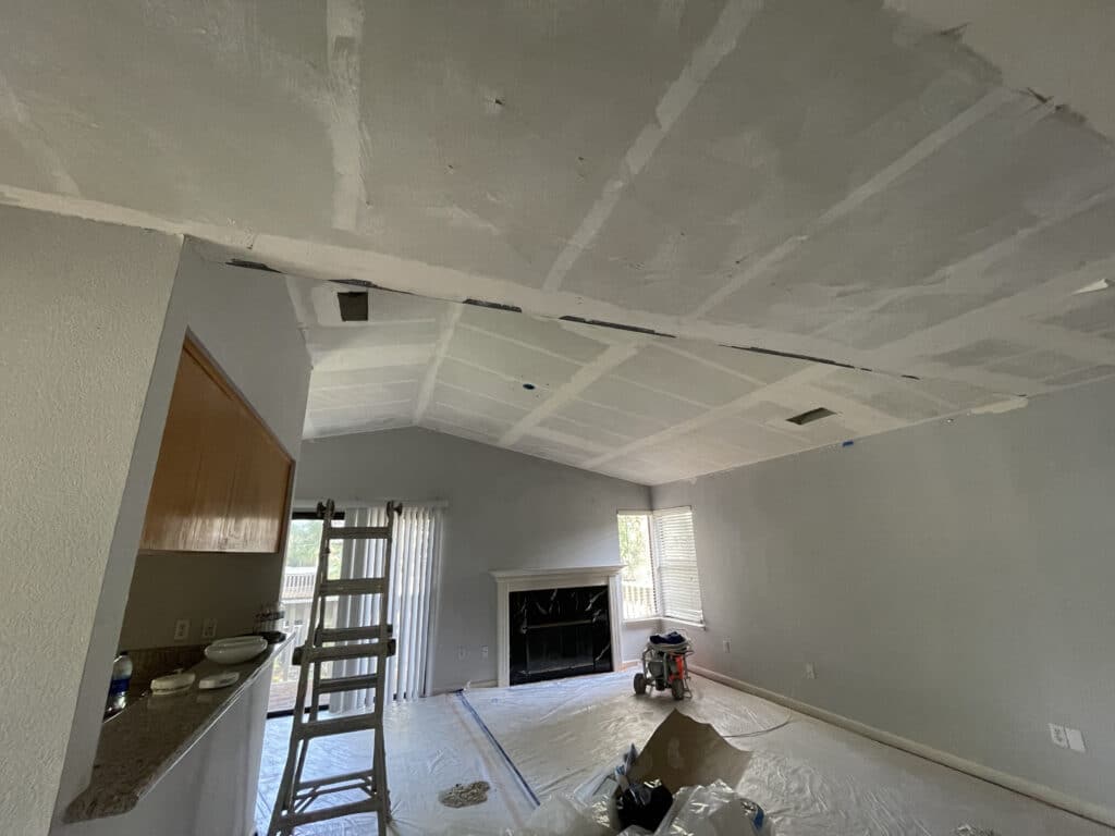 Living room remodel in progress in a Pasadena home, with a vaulted ceiling freshly taped and primed, a ladder, and floors covered in plastic sheeting.