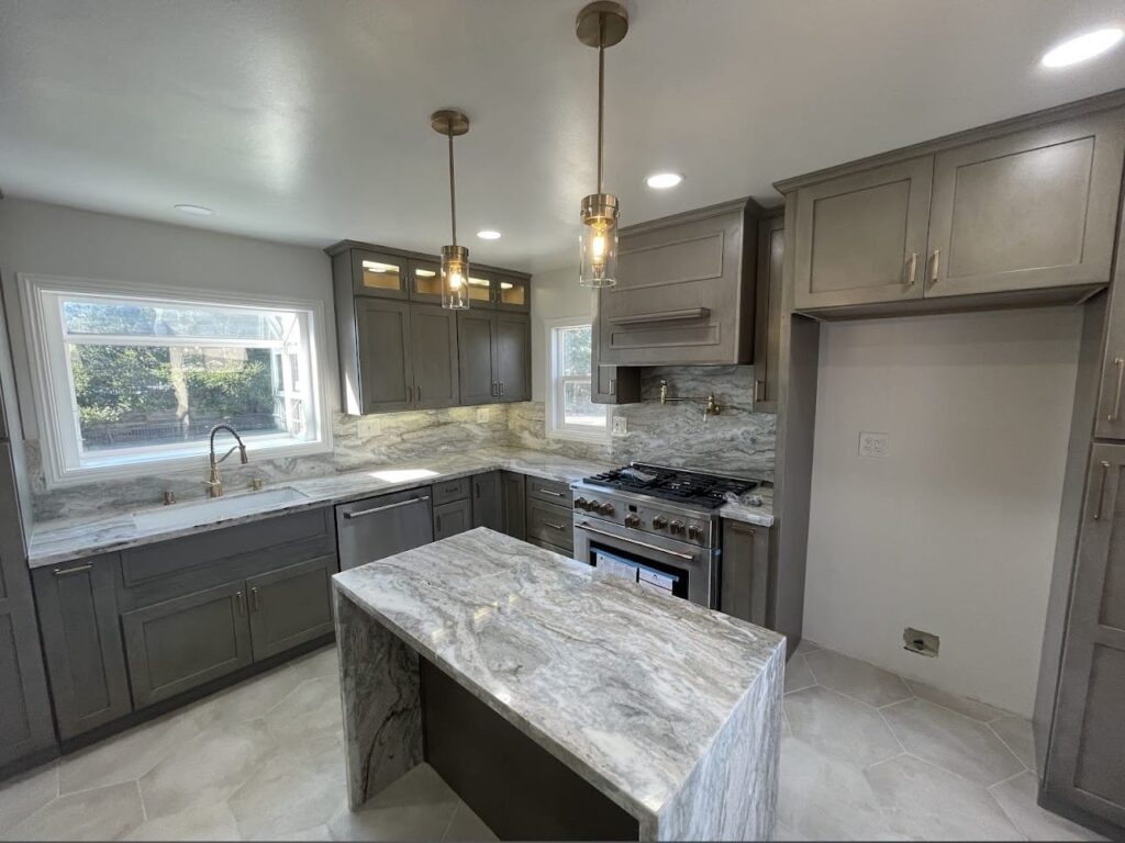 Remodeled kitchen with gray shaker cabinets, white-and-gray marble countertops, a waterfall-edge island, stainless steel gas range, brass pendant lights, and large windows above the sink.