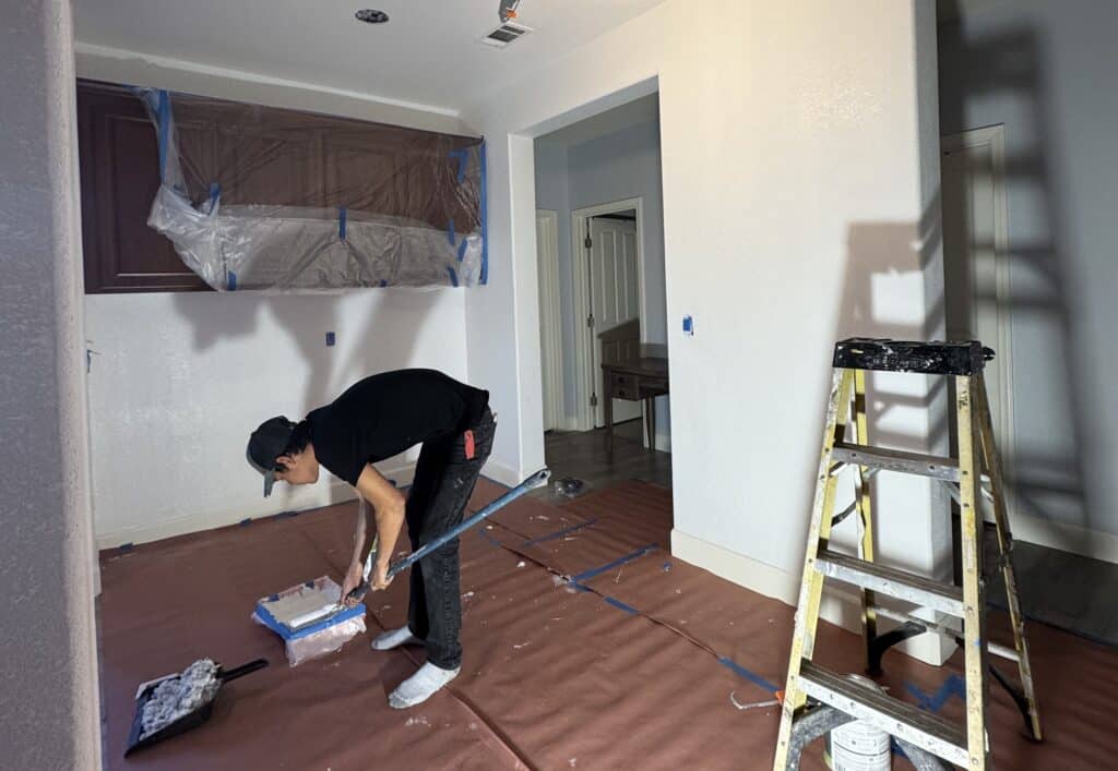 Painter loading a roller from a paint tray in a room prepped for painting, with cabinets wrapped in plastic, paper-covered floors and a ladder nearby.