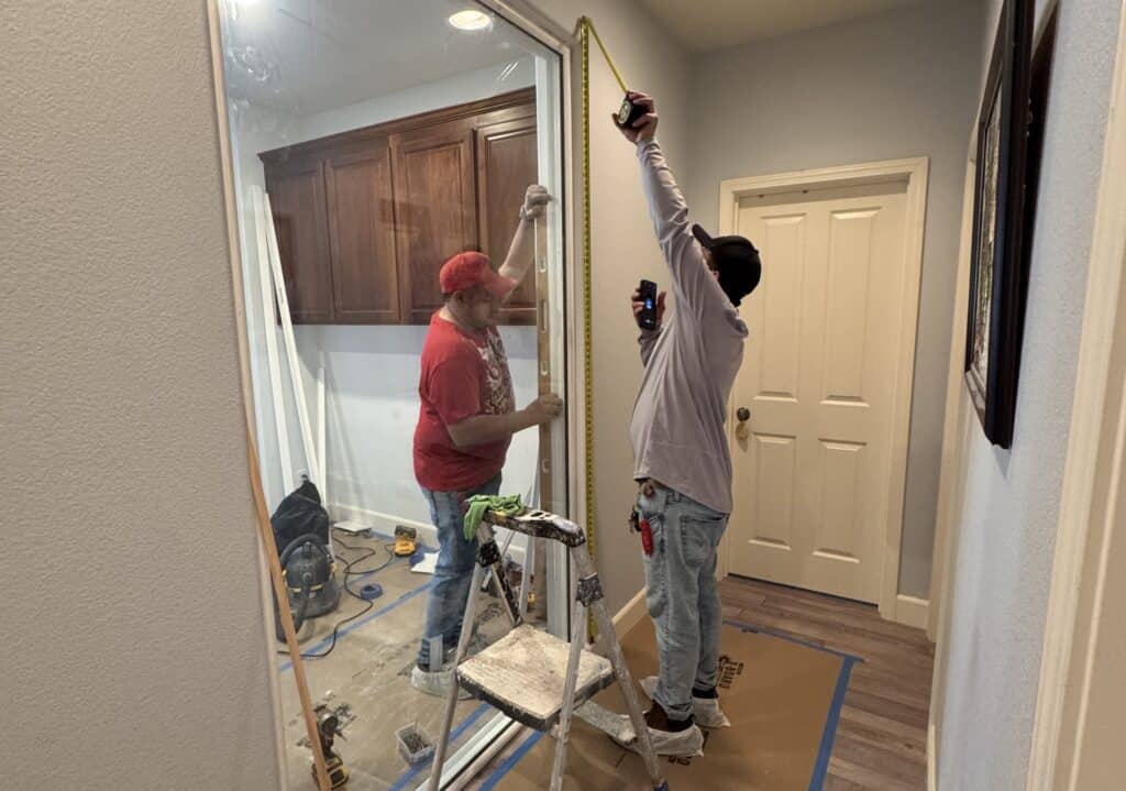 Two workers installing a glass door in a hallway, one checking the frame with a level while the other measures the height with a tape measure, with a step ladder, tools and floor protection around them.